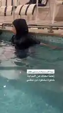 This video demonstrates a woman performing swimming exercises in a pool while wearing a black hijab and swimwear. She begins by standing in the water with her arms raised above her head, then lowers them into the water, creating splashes. The setting appears to be an indoor pool area with lounge chairs visible in the background. Arabic text overlay suggests tips for swimming safely in a hijab. by @activecircle with Original Sound by activecircle