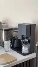 A sleek black coffee machine dispenses hot water into a glass carafe with a paper filter. Next to it, a black kettle sits on its heating base, and a decorative crystal glass cup emits steam, suggesting freshly brewed coffee. The scene captures the essence of a contemporary home coffee station. by @alysf90 with Original Sound by alysf90