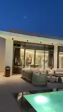 This video captures a serene evening view of a luxurious modern home's outdoor living space. A large, illuminated swimming pool takes center stage, reflecting the deep blue twilight sky. Adjacent to the pool is a spacious patio furnished with elegant, light-colored lounge seating and coffee tables, creating a perfect area for relaxation. Large glass doors and windows reveal the warmly lit interior of the house, showcasing its contemporary design. The overall ambiance is one of tranquility and sophisticated leisure. by @moneeragh with Original Sound by moneeragh