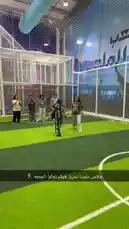 A group of children enjoy playing soccer indoors on a green artificial turf field. They run around, interact with each other, and participate in a game near a small goalpost. The facility features white netting walls, a blue mural with Arabic text, and bright lighting, creating a vibrant and energetic atmosphere. by @gioco_23 with Original Sound by gioco_23