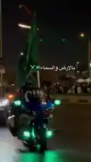 A vibrant night parade celebrating Saudi Arabia's national day. The scene features a motorcycle with glowing green lights leading a procession, followed by people waving large Saudi flags. A spectacular aerial display shows a paraglider trailing bright sparks across the dark sky. The celebration takes place along a decorated street with illuminated arches and crowds of people in traditional attire watching from the sides. by @wedad_23g with Original Sound by wedad_23g