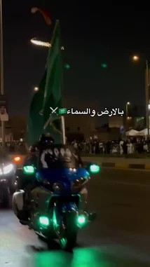 A vibrant night parade celebrating Saudi Arabia's national day. The scene features a motorcycle with glowing green lights leading a procession, followed by people waving large Saudi flags. A spectacular aerial display shows a paraglider trailing bright sparks across the dark sky. The celebration takes place along a decorated street with illuminated arches and crowds of people in traditional attire watching from the sides. by @wedad_23g with Original Sound by wedad_23g