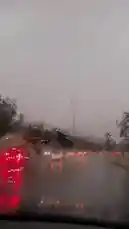 A first-person perspective from inside a car driving on a wet highway during a heavy rainstorm. The windshield wipers are actively clearing the rain, providing intermittent views of the road ahead, other vehicles with their red taillights illuminated, and a large blue overhead highway sign in the distance. The sky is completely overcast and grey, creating a moody atmosphere. by @s3432x with براد الذعاذيع by هادي بن جابر
