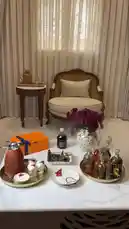 A beautifully arranged living room scene featuring a coffee ceremony setup with ornate trays, a vibrant orange chocolate box, and a vase of deep red flowers. A classic wicker armchair sits beside a small table, creating a cozy and inviting atmosphere. An Arabic poem plays over the scene, adding a layer of cultural richness. by @bee.8907 with Original Sound by bee.8907