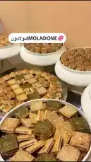This captivating video offers a close-up tour of an impressive array of traditional Arabic pastries, presented by the creator 'مولادون | MOLADONE'. The camera slowly pans across multiple large white bowls and trays filled with an assortment of golden-brown treats. We see delicate, flower-shaped cookies, square sesame bars, and other layered confections, all meticulously arranged. The warm lighting highlights the textures and inviting appearance of these homemade sweets, suggesting a celebration or a special occasion. by @moladone with Original Sound by Contains: Ensan Thani - Adam