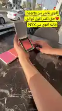 This beauty-focused video features a close-up demonstration of various blush products. The creator applies different shades—ranging from soft pinks and corals to vibrant reds—directly onto their forearm to showcase the pigmentation and finish. The setting appears to be a well-lit indoor space, possibly a boutique or studio, with a view of a street and cars outside. As the swatches are applied, a man's voice sings a romantic Arabic song in the background, creating a relaxed and stylish atmosphere. by @butik.mariya with وجَلست أنـاظرك 💙💙💙. by ً