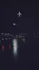 A dark, rainy night scene at an airport tarmac or road. Wet pavement reflects the bright lights of moving vehicles and stationary equipment. A white airplane icon is overlaid at the top center of the frame, suggesting air travel or flight status. The atmosphere is moody and cinematic, capturing the quiet energy of an active airport after dark. by @wolfzzz3 with Original Sound by wolfzzz3