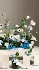 A close-up showcase of beautifully arranged wedding centerpieces and place settings. The display features white calla lilies, blue and white flowers, and lush greenery set against a clean white backdrop. A prominent blue name sign reading 'ABDULAZIZ' adds a personalized touch. The camera pans across elegant black vases holding individual floral arrangements and delicate paper place cards adorned with pearls. This sophisticated setup is perfect for weddings, receptions, or special events. by @r200an with original sound