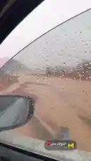 من داخل سيارة، يُظهر المشهد المطر يهطل على طريق ترابي في منطقة صحراوية ذات مناظر طبيعية خلابة. تظهر أشجار قليلة، جبال في الخلفية، وسائقو سيارات متوقفون على جانب الطريق. يعكس المرآة الجانبية جزءاً من السيارة، بينما تغطي قطرات المطر الزجاج الأمامي. يُظهر النص على الشاشة 'إشوڤك مطر ❤️'، مما يشير إلى احتفال أو ترحيب بقدوم المطر في هذه المنطقة. يُعد هذا المشهد فريدًا لكونه يجمع بين الطبيعة الصحراوية والمطر النادر، مما يخلق جواً ساحراً وجميلاً. by @karim.alsaleh with اشوفك مطر♥️ by ا