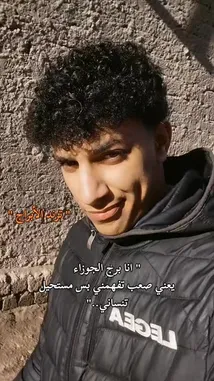This short-form video features the creator, سُوفــــــا Wildcats, against a rustic stone wall. He wears a black puffer jacket with 'LEGEA' branding, looking directly into the camera with a subtle smile. Arabic text overlays his image, conveying a personal message about self-acceptance and resilience. The bright sunlight creates strong shadows, highlighting his curly hair and determined expression, suggesting a narrative of overcoming challenges. by @sofaa_66 with sofaa_66 by Original Sound
