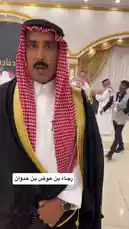 In this close-up shot, Khalifa Eid is seen wearing traditional Saudi clothing, including a white thobe, black bisht with gold trim, and a red-and-white checkered ghutra secured with an agal. He stands in a formal indoor setting, possibly a government building or event hall, with other men in similar attire and national flags visible in the background. His animated expression and direct gaze suggest he is speaking passionately, likely addressing an audience or making a statement related to Saudi culture, heritage, or national pride. by @kh-p19 with kh-p19 by Original Sound