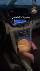 This video provides a close-up view from inside a car, focusing on the center console area. A person holds a glass of coffee, and the camera pans down to show the gear shift, cup holders, and various controls. The interior features beige leather seats, one of which has visible stains. On-screen text in Arabic reads 'التغريدات: سلبيات' (Tweets: Negatives). An animated sticker of a dog with its mouth open is overlaid on the screen, accompanied by the text 'اد العفاريت' (Ouch, devils!). The scene captures a casual, everyday moment. by @kfl1 with Original Sound by kfl1
