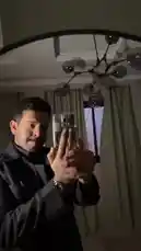 In a dimly lit room, Abduallah Tale demonstrates his personal style and filming technique. He holds a smartphone, adjusting its position to capture his reflection in a large mirror. The modern, dark-colored chandelier above him adds a touch of elegance to the setting. This scene offers viewers a behind-the-scenes look at how he prepares and films his content. by @r011.z with Original Sound by r011.z