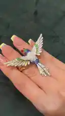 A close-up showcase of a stunning, multi-colored hummingbird brooch. The piece is first displayed on a hand, then elegantly transformed into a floral cuff bracelet, demonstrating its intricate design and wearable versatility. by @moneeragh with Original Sound by moneeragh