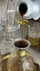 This elegant video captures a serene ritual centered around luxury fragrances and incense. It begins with dark perfume being poured into a crystal bottle using a gold-topped funnel. The scene transitions to a hand in a white glove lighting a glowing charcoal disc with a brass torch, which is then placed into a traditional wooden incense burner. The final shots reveal a luxurious collection of agarwood chips, various perfumes, and other aromatic items arranged on ornate trays, all set against a backdrop of soft smoke, creating a sophisticated and tranquil atmosphere. by @bee.8907 with Original Sound by bee.8907
