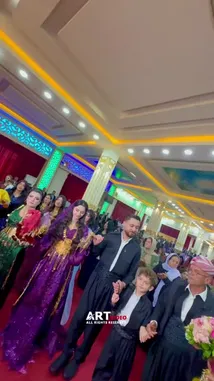 This vibrant scene captures a traditional cultural celebration featuring a group of people dressed in elaborate, colorful attire performing a synchronized dance. The setting is an ornately decorated hall with intricate ceiling designs and warm lighting, suggesting a festive occasion such as a wedding or festival. Participants, including men, women, and children, clap rhythmically and move gracefully, embodying joy and community spirit. The atmosphere is lively and celebratory, highlighting the beauty of shared traditions. by @artvideo.me with artvideo.me by Original Sound