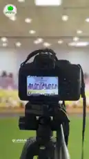 This video offers a behind-the-scenes look at a school performance being recorded by a professional camera on a tripod. On the camera's screen, a group of young performers in matching purple dresses can be seen on stage, singing a song in Arabic. The setting appears to be a large hall or auditorium within the Al-Faisal Private Schools, capturing a moment of student talent and achievement. by @ajyalf with Original Sound by ajyalf