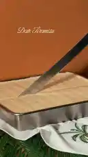 A person dressed in traditional Middle Eastern attire performs an impressive act of cutting a rectangular 'Date Tiramisu' cake with a large, ornate sword. The dramatic reveal shows the layered cake on a plate, demonstrating a unique and visually striking method of serving a dessert. by @fox_cofee with Original Sound by fox_cofee