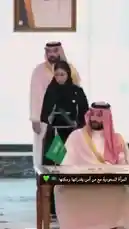 A man in traditional Saudi attire, wearing a white thobe and red-and-white checkered ghutra, sits at a wooden desk signing official documents. A small green Saudi flag with Arabic script stands prominently on the desk. Behind him, another man in similar regalia observes, while a woman in a black abaya holds a folder. The setting appears formal, suggesting a significant governmental or state event. by @batoolmakeup1 with Original Sound by batoolmakeup1