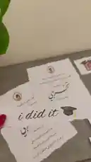A close-up view of personalized graduation cards written in Arabic script. Each card features a photo of a graduate, a congratulatory message, and a graduation cap icon, all laid out on a table. The scene evokes a sense of achievement and celebration. by @rubaaomaar with Original Sound by rubaaomaar