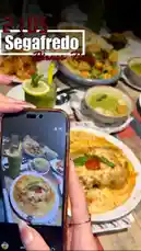 A close-up shot shows a hand holding a smartphone, capturing a vibrant dinner spread at Segafredo. The table is filled with colorful dishes like a cheesy baked dish topped with tomato and basil, a bowl of green soup, and a refreshing lemon drink. A text overlay reads 'Segafredo Dinner Time' with the time '21:05'. This scene perfectly illustrates the art of photographing food in a restaurant setting. by @segafredohebron with original sound