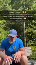 A man with a beard and tattoos sits on a wooden bench in a lush green forest, speaking directly to the camera. He greets viewers with 'Good Wednesday,' mentions the sunny weather, and discusses plans to visit his workshop where he'll receive a new compressor. He then shifts the topic to a philosophical discussion about democracy, questioning its true meaning and application. by @vikingcustom with Original Sound by vikingcustom