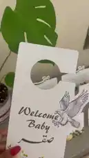 A close-up showcase of elegant 'Welcome Baby' door hangers and matching Arabic baby shower cards from the brand قرطاسية سما. The items feature a beautiful eagle illustration and floral designs. A baby can be heard crying softly in the background, adding a realistic touch to the celebratory theme. by @rubaaomaar with Original Sound by rubaaomaar