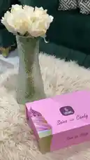 This video provides a full unboxing and close-up look at the SOINS 'Soin Ice Candy' perfume set. A hand opens the elegant pink box to reveal a larger Eau de Parfum bottle and a smaller travel-sized bottle, showcasing their matching labels and packaging. The bottles are displayed side-by-side on a fluffy white rug next to a vase of white roses, highlighting the product's aesthetic appeal. by @soins_sa with Original Sound by soins_sa