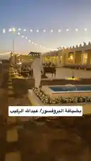A serene evening at a traditional Saudi venue, where men in white thobes and ghutras stand in formation. String lights illuminate the courtyard as a group chants together in unison, creating a peaceful and culturally rich atmosphere. by @y_alkhmsan with Original Sound by y_alkhmsan