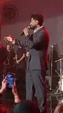 Experience the electrifying atmosphere of Mohamed Fouad's live concert. This video captures the singer in a sharp suit, passionately performing on stage under vibrant red lights. He engages with his band, including guitarists and keyboardists, while the enthusiastic crowd cheers and records the moment on their phones. The performance is a dynamic showcase of music and connection. by @mo.fouadziko with Original Sound by mo.fouadziko