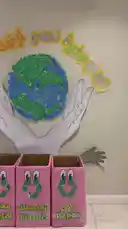 This educational video shows children at the 'Step by Step' child care center actively participating in a hands-on recycling lesson. They sort various types of waste into labeled pink bins for metal, plastic, and paper, guided by a large, colorful Earth-themed display on the wall. The children work together to clean up and organize the materials, demonstrating teamwork and environmental responsibility in a fun and interactive classroom setting. by @stepbystepkh with Original Sound by stepbystepkh