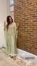 A woman showcases a beautiful light green kaftan dress with intricate floral embroidery. She turns gracefully against a rustic brick wall, highlighting the garment's flowing silhouette and detailed craftsmanship. This look offers inspiration for elegant, modest fashion styling. by @needle_t with موسيقى عود - الجمال by اسامه عبدالهادي