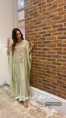 A woman showcases a beautiful light green kaftan dress with intricate floral embroidery. She turns gracefully against a rustic brick wall, highlighting the garment's flowing silhouette and detailed craftsmanship. This look offers inspiration for elegant, modest fashion styling. by @needle_t with موسيقى عود - الجمال by اسامه عبدالهادي