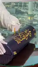 A woman in traditional white lace attire unboxes a luxurious dark blue crocodile-textured prayer mat case with gold Arabic calligraphy. She reveals the rolled-up navy blue prayer mat with golden crescent and star embroidery, then sets it up on the carpeted floor beside a large window overlooking the Kaaba in Mecca. She kneels to pray, using a string of prayer beads, showcasing a serene moment of devotion. by @joy.narcissus with Original Sound by joy.narcissus