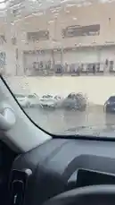 A view from inside a car during a rainy day, showing water droplets on the windshield. A white SUV is seen driving alongside. An Arabic speaker comments on the weather, saying 'The world has flooded, Mashallah,' while expressing concern about getting out. The scene captures the experience of navigating through heavy rain. by @guruofmarketing with Original Sound by guruofmarketing