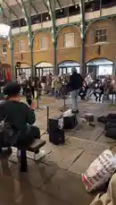 A street musician performs for an engaged audience seated on benches inside a historic covered market hall. The performer, holding a microphone, sings into a stand while a small crowd watches and records the moment on their phones. The venue features distinctive arched brick walls and a high, vaulted ceiling with metal trusses and hanging lights, creating a warm and atmospheric setting for the impromptu concert. by @moneeragh with Original Sound by moneeragh