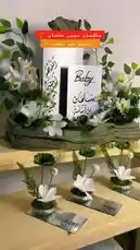 Watch this elegant display of baby shower decorations perfect for a traditional celebration. The centerpiece features beautiful white flowers, lush greenery, and custom Arabic calligraphy cards reading 'Baby' and 'مرحباً بالطفل'. Below, four matching table centerpieces are arranged neatly. This video is ideal for anyone seeking inspiration for Arabic baby shower decor. by @r200an with original sound