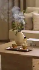 A serene, slow-motion shot of a beautifully styled coffee table in a cozy living room. The scene features a textured vase with delicate green branches, a small incense burner releasing a gentle plume of white smoke, two colorful espresso cups on a tray, and books stacked nearby. In the background, soft-focused beige sofas create a warm atmosphere. This tranquil setting is accompanied by the sound of a man reciting Arabic, enhancing the peaceful and meditative mood. by @om-5p with Original Sound by om-5p