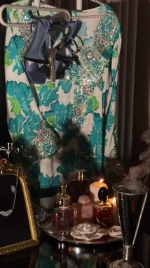 A stylish ensemble is displayed, featuring a vibrant floral dress adorned with sparkling embellishments, paired with elegant dark blue strappy heels. The scene is completed with a curated collection of accessories, including a delicate necklace, perfume bottles, and a lit candle, creating a sophisticated and glamorous atmosphere. by @ab.7955 with Original Sound by ab.7955