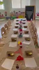 This lively video captures young children at the 'Step by Step' child care center participating in a hands-on pizza-making activity. Dressed in playful chef hats, the kids are seated at a long table set with ingredients like bread slices, colorful toppings in paper boats, and condiment bottles. The atmosphere is cheerful and educational, with close-ups showing the children's excitement and engagement as they prepare their individual creations. by @stepbystepkh with Original Sound by Contains: Pizza Party - Super Simple Songs, Caitie's Classroom
