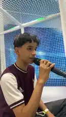 A young boy with curly hair, wearing a maroon and white sports jersey, sits in a modern play area with blue tiled walls and white netting. He holds a black microphone close to his mouth, appearing to sing or speak with great focus and expression. The background features a vibrant blue pattern, suggesting a dynamic and youthful environment. by @gioco_23 with Original Sound by gioco_23