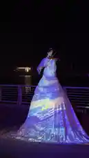 A woman performs on a violin at night on a waterfront promenade. She wears a long, white gown with a voluminous skirt, which is illuminated by a dynamic light projection showing swirling patterns of purple, blue, yellow, and orange. As she plays, she turns slightly, revealing a large, colorful 'WONDER HILL' sign in the background. The performance is artistic and visually captivating. by @home_garden3 with Original Sound by home_garden3