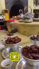 This video showcases a beautifully arranged traditional Arabic tea setup. The scene features multiple silver bowls filled with dates placed on a counter in front of a glowing fireplace. Two shiny metal teapots sit ready for use, while shelves above display various decorative items. A red-lidded jar, a small brush, and a box labeled 'دم البدود' (a type of spice) are visible, adding to the authentic atmosphere. The warm firelight creates a cozy ambiance, perfect for serving dates and tea. by @s3432x with Original Sound by s3432x