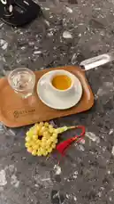 This close-up shot captures a serene coffee moment. A hand uses a wooden spoon to gently stir espresso in a white cup, which rests on a wooden tray branded 'STEAM ROASTERS'. Next to the coffee is a glass of water and a distinctive yellow prayer bead necklace with red tassels. The entire scene is set on a dark, textured marble countertop, creating a calm and contemplative atmosphere. by @alysf90 with Original Sound by alysf90