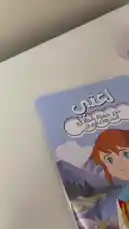 Watch as Ruba from 'قرطاسية سما' unboxes and reviews a beautiful spiral-bound notebook featuring a character from the animated series 'LGBTI'. The notebook has a charming cover design with mountains, flowers, and the Arabic text 'لإغتي حور حمزة باحاتي' and 'الاسم الثالث الأسود'. She flips through the pages, showing the lined paper and decorative borders, before placing it back on her desk next to a pink calendar. by @rubaaomaar with Original Sound by rubaaomaar