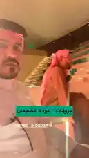The video features Yaser Al-Khamsan, dressed in traditional Saudi clothing including a thobe and a red-and-white checkered ghutra, attending an event inside a large, modern auditorium. He is seated among other men in similar attire, listening attentively to a speaker who is visible on a stage in the background. The setting has tiered seating with teal-colored seats, and the atmosphere suggests a formal gathering such as a conference or lecture. by @y_alkhmsan with Original Sound by y_alkhmsan