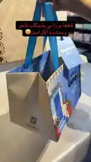 This short clip provides a detailed look at a chocolate product. It begins with a hand holding a blue gift bag featuring 'Guyen' branding and Arabic text 'برواني بيشوكت فاخر وبحشوة الكراميل' (Premium Brownie Chocolate with Caramel Filling). The bag is opened to reveal a tin box with a coastal scene. Inside the tin, neatly arranged square brownie chocolate bars are shown. A person wearing gloves breaks one bar open, revealing its rich, dark brownie base and a gooey caramel filling. The video serves as a visual review and unboxing of these premium chocolate treats. by @ch11.30 with Original Sound by Contains: Pretty Little Baby - Connie Francis
