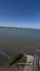 This tranquil scene captures a beautiful coastal landscape under a clear blue sky. The camera pans slowly across calm waters towards a distant shoreline dotted with buildings and structures, creating a peaceful atmosphere. by @tr3i5 with وانا في حاله غريبه by .