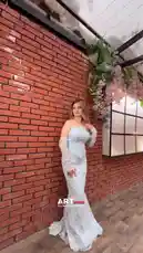 A woman poses gracefully in a stunning light blue lace mermaid gown against a rustic red brick wall adorned with lush greenery and pink flowers. She wears matching long lace gloves and strikes various elegant poses, showcasing the intricate details of her strapless dress. The setting features modern decor with a large illuminated panel, creating a chic and romantic atmosphere perfect for a photoshoot. by @artvideo.me with artvideo.me by Original Sound