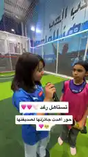 This dynamic footage captures a lively moment on an indoor soccer field. A young girl in a blue Puma jersey enthusiastically interviews another player wearing a pink bib, holding a microphone. The setting is a modern sports facility with artificial turf, white safety netting, and Arabic script on the walls. The atmosphere is energetic, suggesting a youth tournament or training session. by @gioco_23 with Original Sound by gioco_23
